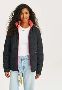 Prošívaná černá bunda s růžovým límcem a zipem, se barevným třásněm na táhle zipu. Nositá přes bílé tričko.