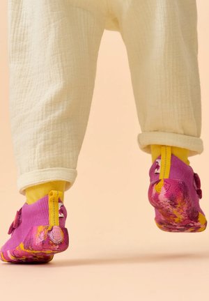 Bambino che indossa pantaloni crema, calzini gialli e scarpe viola per bambini con suola gialla, che cammina su una superficie beige.