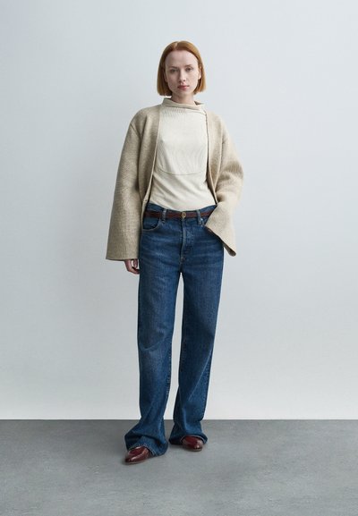 Μπεζ μεγάλο cardigan, κρεμ εφαρμοστό τοπ, μπλε τζιν με φαρδιά μπατζάκια, καφέ ζώνη με κυκλική αγκράφα και καφέ παπούτσια. Απλό φόντο.