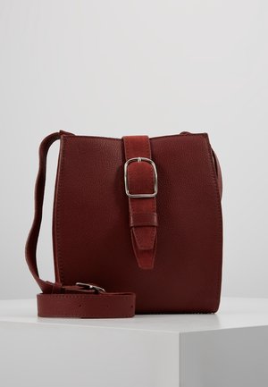 Tummanpunainen nahkainen crossbody-laukku, jossa säädettävä hihna ja keskellä mokkanahkainen solkikoriste, valkoisella jalustalla esillä.