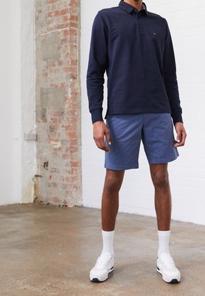 Hombre vestido con un polo de manga larga color marino, pantalones cortos azules, calcetines blancos y zapatillas blancas, de pie en un interior sobre un suelo de hormigón al lado de una pared de ladrillo.