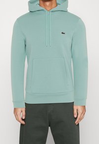 Felpa leggera color teal con tasca frontale. Presenta lacci di regolazione e un piccolo logo ricamato. Realizzata in un tessuto morbido e testurizzato. Vestibilità casual.