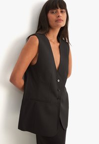 Musthave musta ilma varrukatega vest sügava V-kaelusega, ühekohaline esitlus ja kaks taskut. Valmistatud siledast kangast, millel on kohandatud lõige.