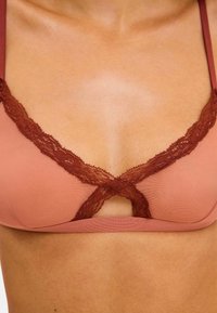 Vaaleanpunainen rintaliivi, jossa on tummemman punaruskeat pitsireunat, ja keskellä koristeellinen aukkoasu sekä säädettävät hihnat.