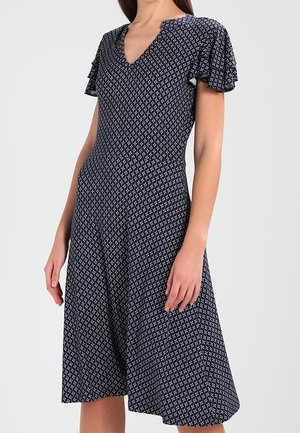 Mujer con vestido azul marino hasta la rodilla, con pequeños patrones geométricos blancos y mangas cortas con volante.