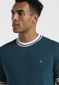 Teal T-shirt met korte mouwen en een witte ronde hals, versierd met gestreepte accenten. Bevat een klein pinguïnlogo op de linkerborst. Heeft een soepele textuur.