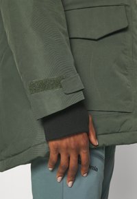 Grüne isolierte Jacke aus strukturiertem Stoff mit einem Klettverschluss am Manschettenbereich, kombiniert mit einem schwarzen Strickbündchen. Die Hand liegt auf einer hellblauen Hose.