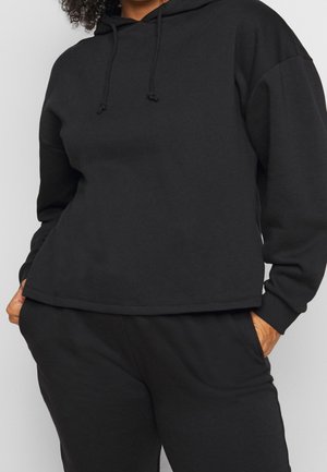 Persoon die een zwarte hoodie en bijpassende zwarte broek draagt met handen in de zakken, toont een casual ontspannen outfit.