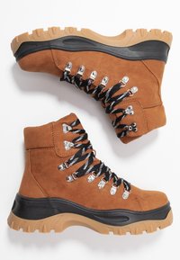 Bottines en daim marron avec des lacets noirs et blancs. Dotées d'œillets métalliques et d'une semelle en caoutchouc beige robuste avec des motifs antidérapants.