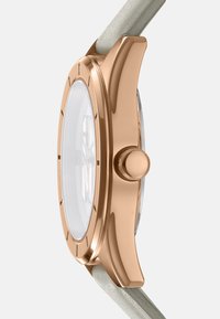 Ronde horloge met een kast van rosegoud roestvrij staal, een heldere glazen wijzerplaat en een lichtgrijze leren band. Beschikt over een centrale kroon aan de zijkant.