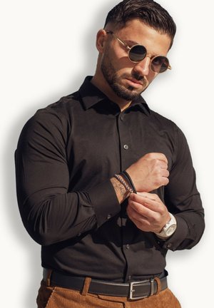 Black button-up shirt with rolled sleeves; wearing silver watch and layered bracelets; textured brown pants; background white.