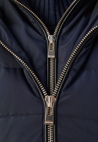 Veste matelassée bleu marine avec deux fermetures éclair symétriques en argent aux dents dorées, coutures texturées et col côtelé.