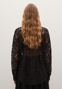 Persona con lunghi capelli castano chiaro mossi indossa una blusa in pizzo floreale nero trasparente, in piedi di spalle contro uno sfondo chiaro e semplice.