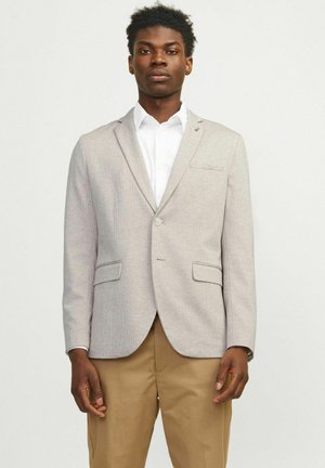 Blazer gris claro con una tela texturizada, cierre de un solo botón, dos bolsillos delanteros y un sutil acento cerca de la solapa. Combinado con una camisa blanca.
