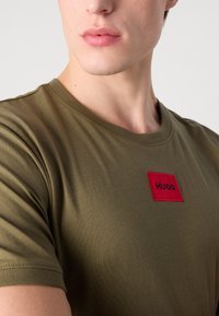 Olívazöld póló puha anyagból, piros téglalap alakú folttal, amelyen vastag fekete betűkkel áll a "HUGO" felirat. Klasszikus kerek nyakú dizájn.