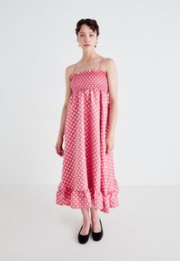 Pink polka dot dress with a smocked bodice, thin straps, and a flared design. The hem features a ruffled accent. Worn with black shoes.