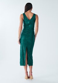 Žalia suknelė su blizgančiomis akutėmis, aptemptu siluetu, V formos nugarėle, petnešėlėmis ir aukšta šlaunies skeltuku. Lygi tekstūra su žėrinčiais akcentais.