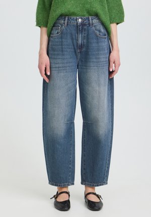 Personne portant un jean bleu délavé droit, un pull vert et des ballerines en cuir noir, debout devant un fond uni.