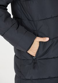 Mustavillainen puhvijakku, jossa näkyy käsi vetoketjullisessa taskussa. Materiaali on sileä, jossa on tikattu pinta ja joustavat hihansuut.
