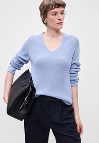 Ανοιχτό μπλε πλεκτό πουλόβερ με V λαιμόκοψη, συγκεντρωμένα μανίκια, συνδυασμένο με σκοτεινές ριγέ παντελόνες και μαύρη δερμάτινη τσάντα.