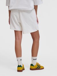 Άτομο που φοράει λευκά σορτς, λευκές κάλτσες Hummel και κίτρινα παπούτσια Hummel με πράσινες λεπτομέρειες, από πίσω.