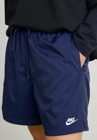 Person som bär marinblå Nike-shorts med vit logotyp, dragsko i midjan, och svart långärmad tröja, hand i höger ficka.