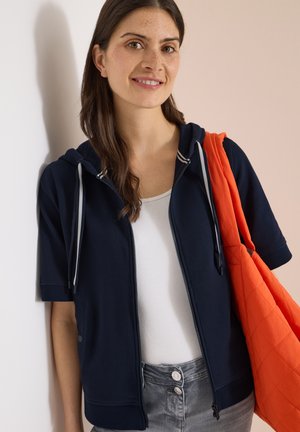 Kobieta o długich brązowych włosach, ubrana w granatową bluzę z krótkim rękawem z kapturem, biały top, szare dżinsy i nosząca na ramieniu pomarańczową pikowaną torbę typu tote.