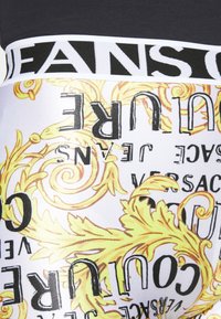 Pás s pevným pasem s nápisem "JEANS" v černé na bílém, kontrastující s vzorovanou tkaninou s ornamentálními zlatými vzory a textem v černé a zlaté.