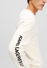 Krémová mikina s okrúhlym výstrihom a čiernym textom "KARL LAGERFELD" potlačeným vertikálne na ľavom rukáve. Mäkká textúra.