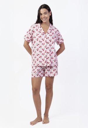 Joven sonriente, vestida con un conjunto de pijama de manga corta a cuadros rosas con estampado de cerezas rojas, de pie descalza contra un fondo blanco.