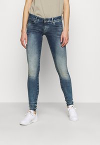 Mujer vestida con jeans ajustados azul desgastados y zapatillas blancas, de pie contra un fondo liso con las manos a los lados.