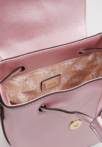 Vaaleanpunainen synteettinen käsilaukku kiiltävällä pinnalla, jossa on kullanvärinen painonappihela ja kuvioitu kangasvuori, merkinnällä "GUESS".