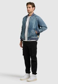 Homme portant une veste bomber en denim bleu, un t-shirt blanc, un pantalon cargo noir et des baskets montantes blanches, debout avec une main dans la poche.