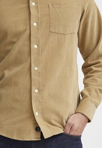 Tan corduroy shirt met een zak, knoopsluiting en een textuur stof. De onderkant is licht gebogen met een logo-label aan de onderzijde.