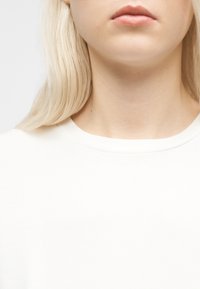 Gros plan sur le bas du visage, le cou et le haut de la poitrine d'une personne à la peau claire, cheveux blonds, portant un t-shirt blanc uni à col rond.