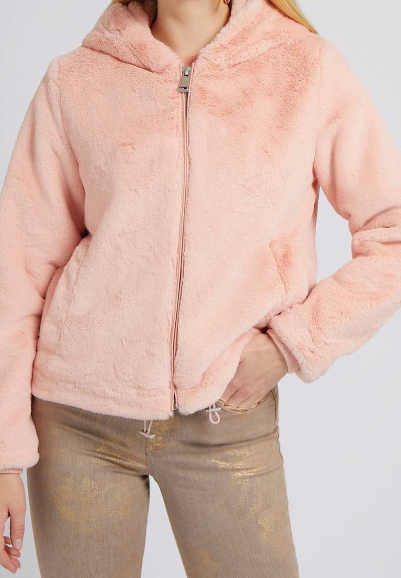 Femme portant une veste zippée duveteuse rose clair avec capuche et un pantalon slim finition or métallisé, une main dans la poche de la veste.