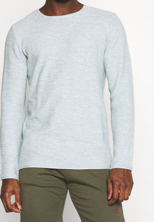 Homme portant une chemise à manches longues gris clair texturée et un pantalon vert olive, debout avec les bras détendus le long du corps.