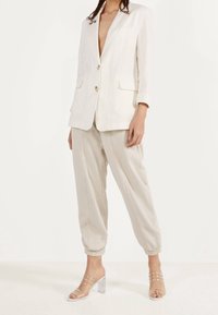 Lněný sako v krémové barvě s klopovými límci a dvěma předními kapsami, doplněné světle béžovými tapered kalhotami a sandály s průhledným podpatkem.