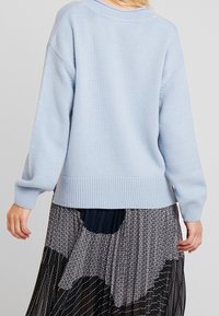 Pull en maille bleu clair avec une coupe décontractée, poignets et ourlet côtelés. Porté par-dessus une jupe à motifs présentant des dessins en noir et blanc.