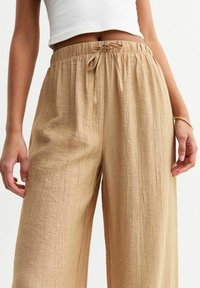 Pantalones de pierna ancha en beige con un acabado texturizado, cintura elástica y detalle de lazo en la parte delantera, fabricados en material ligero.