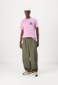 Lyserød bomulds t-shirt med et grafisk patch, parret med løse grønne bukser. Beige sneakers har lyse accenter og en tekstureret sål.