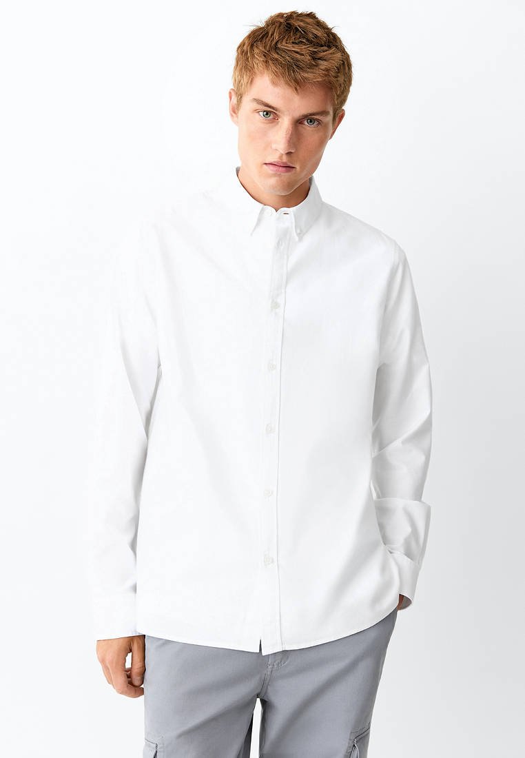 Jeune homme aux cheveux châtain clair portant une chemise blanche boutonnée et un pantalon gris clair, debout sur un fond blanc uni.