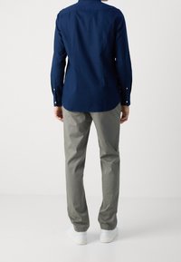 Chemise à manches longues marine, en tissu coton, avec des poignets à boutons, détail d'empiècement au dos ; associée à un pantalon gris clair et des baskets blanches.