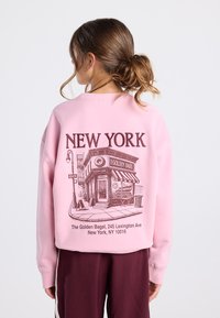 Femme avec les cheveux attachés portant un sweat-shirt rose sur lequel figure un croquis détaillé de la boutique The Golden Bagel et l'adresse de New York au dos.