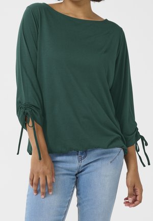 Donna che indossa una maglia a maniche lunghe verde scuro con coulisse regolabili sulle maniche e jeans azzurri su sfondo uniforme.