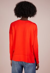 Czerwony sweter z długim rękawem o luźnym kroju, zaokrąglonym dekoltem i rozcięciami po bokach, zestawiony z jasnoniebieskimi dżinsami. Gładka faktura, casualowy design.