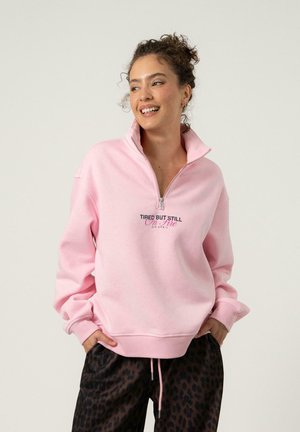 Ljusrosa sweatshirt med halvmoln, ribbade muddar och en grafisk tryck som säger "TRÖTT MEN ÄNDÅ I BRAND" i svart och rosa.