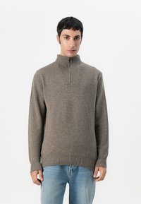 Bruine gebreide trui met een hoge kraag, voorzien van verticale ribbels bij de hals. Afgewerkt met ribbels aan de mouwen en onderkant. Draagt over een spijkerbroek.