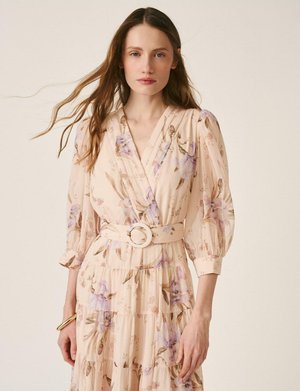 Mujer con cabello largo y castaño, vistiendo un vestido floral beige con mangas abullonadas y un cinturón a juego con hebilla redonda, posando frente a un fondo liso.