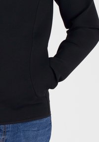 Felpa nera con una texture liscia, caratterizzata da un design aderente e cuciture laterali sottili. Abbinata a jeans in denim blu in vita.
